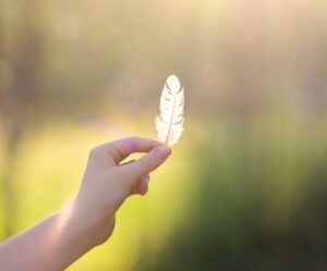 A delicate feather is held in a hand with glowing sunlight in the background.